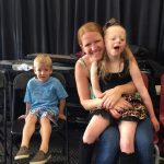 A woman happily embraces a smiling young girl, while a boy sits nearby.