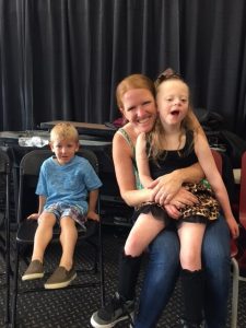 A woman happily embraces a smiling young girl, while a boy sits nearby.