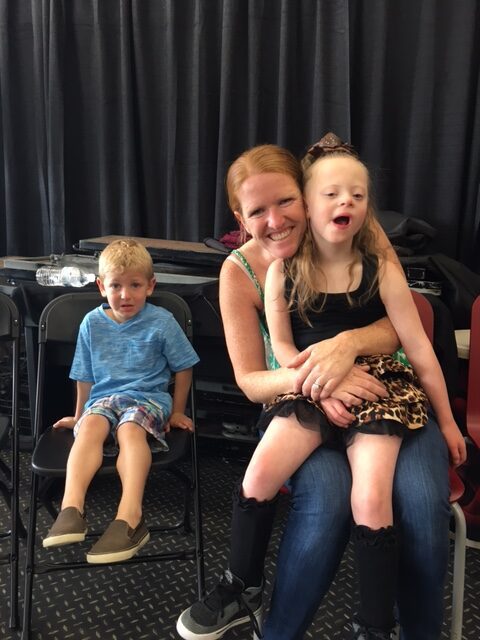 A woman happily embraces a smiling young girl, while a boy sits nearby.