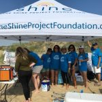 Volunteers in blue shirts under a Shine Project tent at the beach.