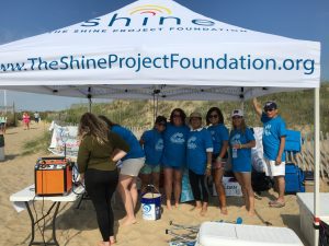 Volunteers in blue shirts under a Shine Project tent at the beach.