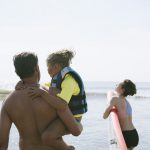 A father carrying his child in a life jacket at the beach.