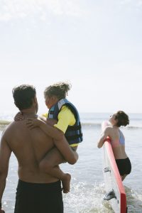 A father carrying his child in a life jacket at the beach.