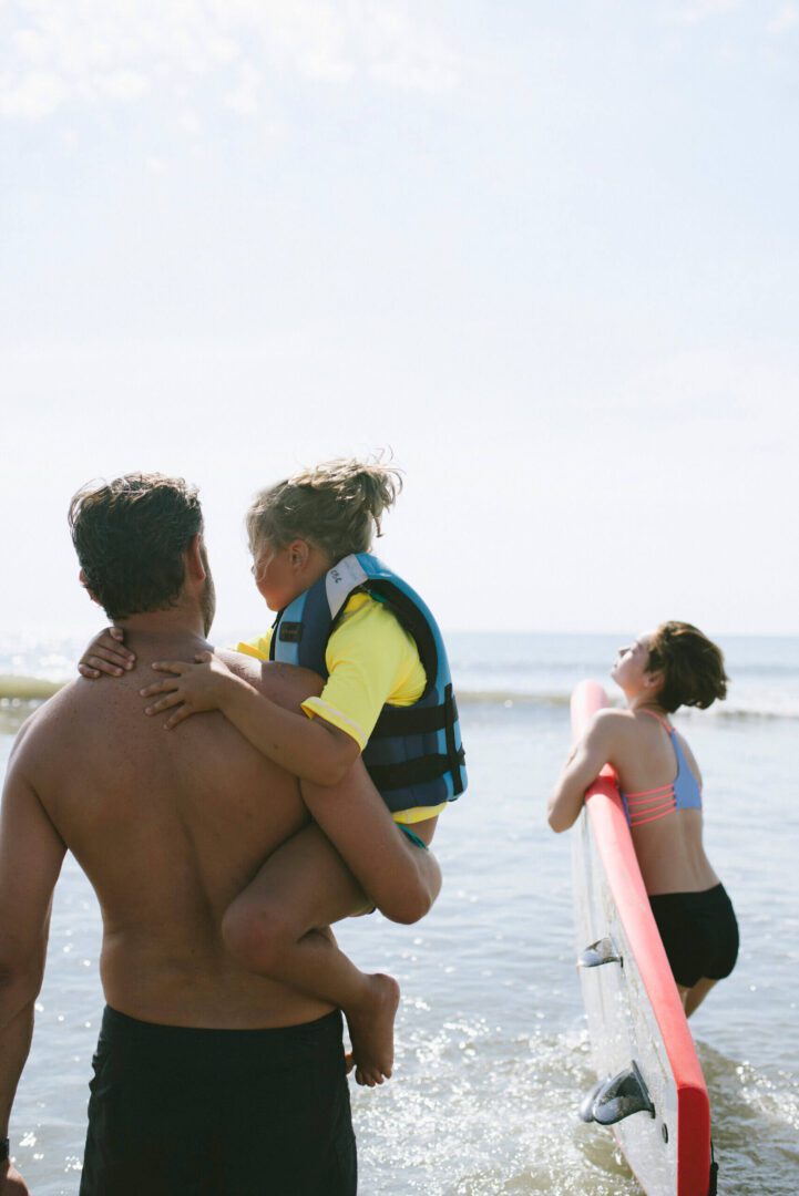 A father carrying his child in a life jacket at the beach.