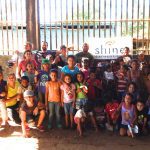 Group of smiling children and adults posing outdoors by a lake with a banner.