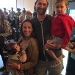 A happy family posing indoors with their two children, one in a stroller and the other held in arms.