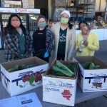 Four women standing behind boxes filled with fresh produce in a market setting.