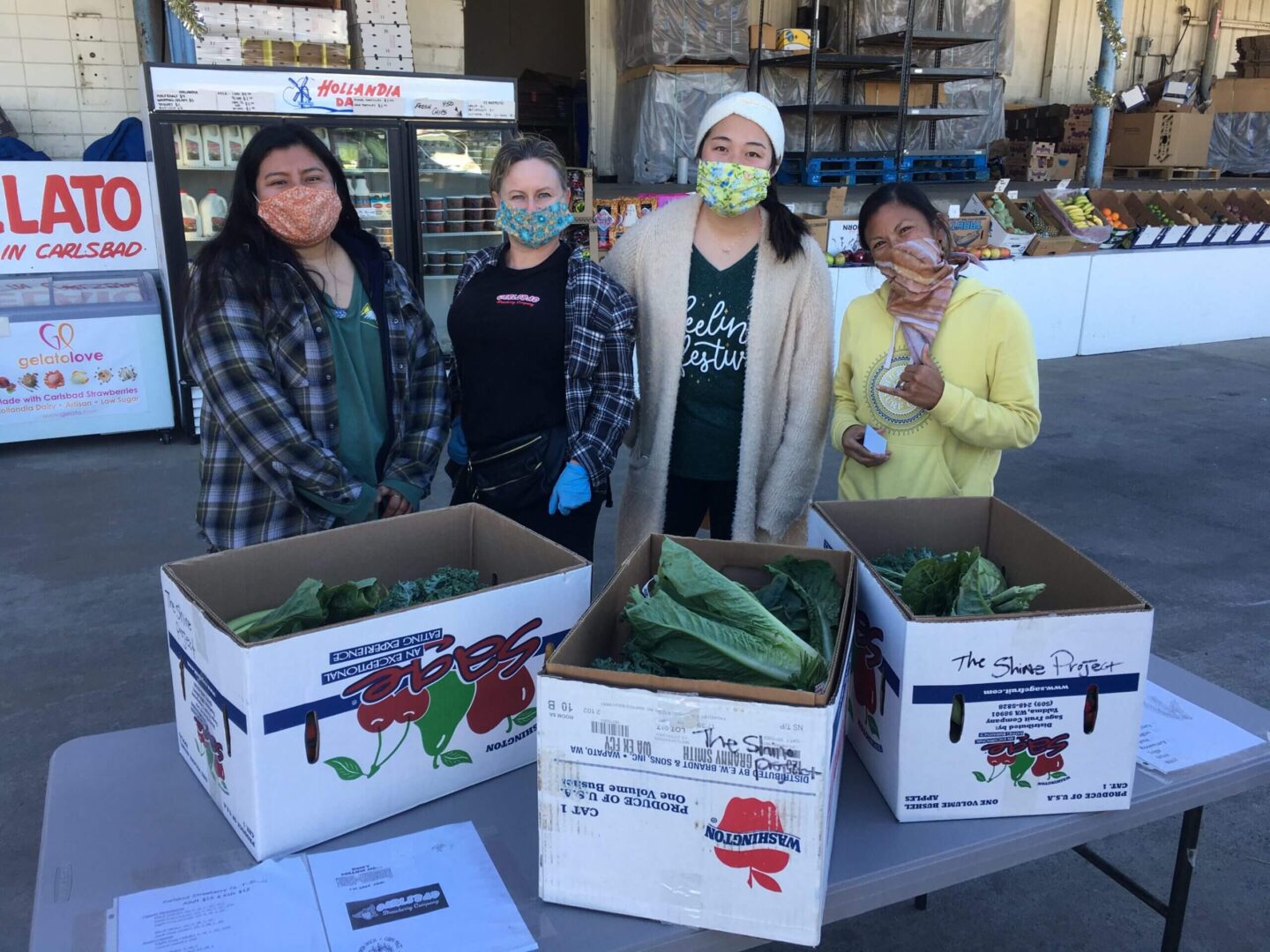 Four women standing behind boxes filled with fresh produce in a market setting.