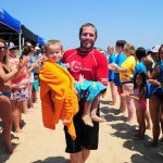 Man carrying a child wrapped in a towel on a crowded beach.