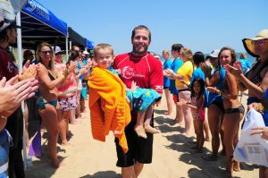 Man carrying a child wrapped in a towel on a crowded beach.