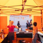 People gather under a bright orange tent at an outdoor event.