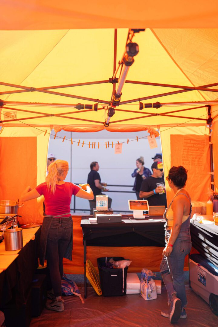 People gather under a bright orange tent at an outdoor event.