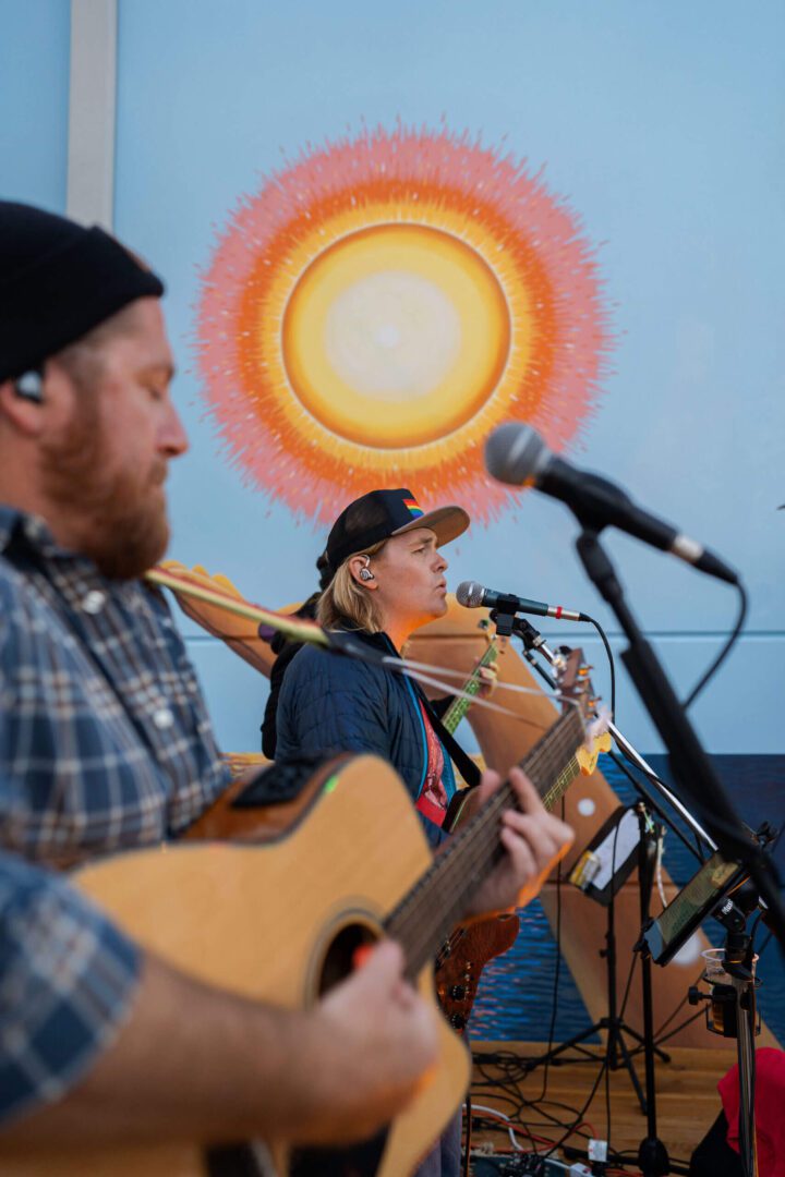 Musicians play guitar and saxophone with vibrant sun mural background.