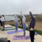 People practicing yoga outdoors on mats near a foggy lake.
