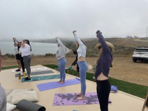 People practicing yoga outdoors on mats near a foggy lake.