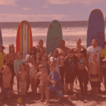 Group of people posing with surfboards on a beach at sunset.