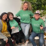 Four smiling adults sitting and standing outdoors, two wearing green AMC t-shirts.