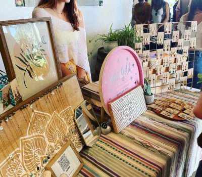 A woman stands behind a table displaying handcrafted wooden and fabric items at a market.