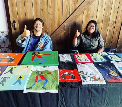 Two people showing peace signs behind a table with colorful artwork.