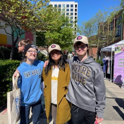 Three friends posing outdoors on a sunny day with trees and buildings in the background.